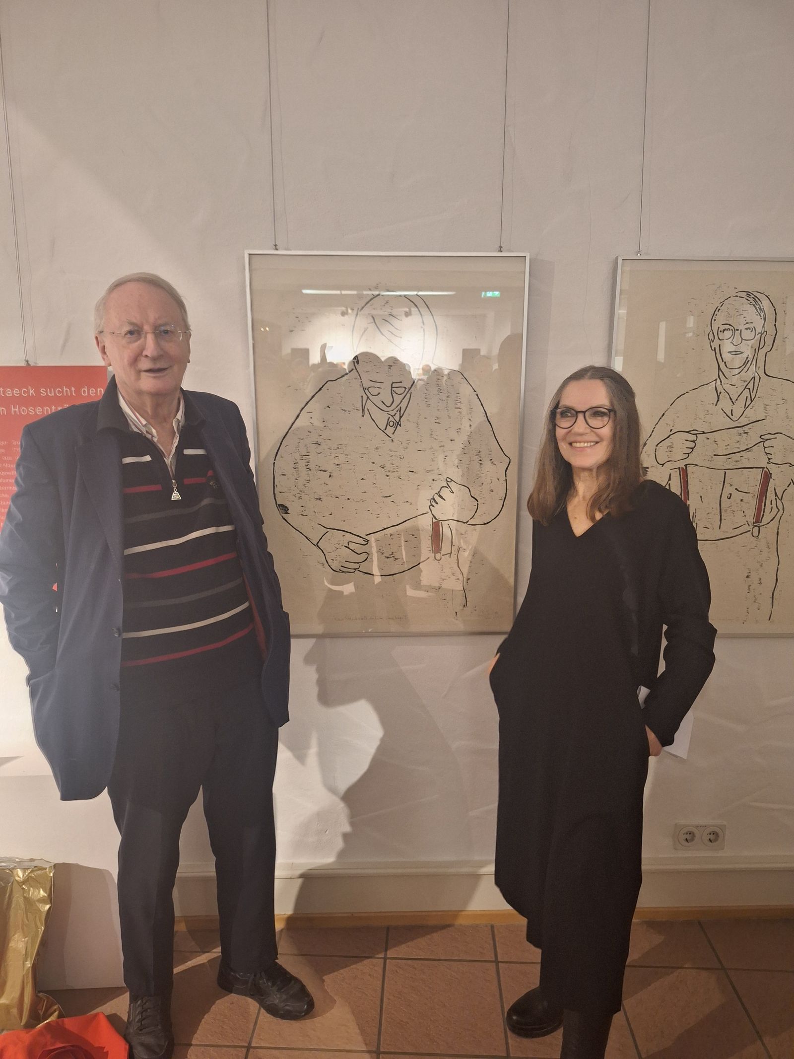 Zwei Personen stehen in einem Ausstellungsraum neben gerahmten Zeichnungen.
