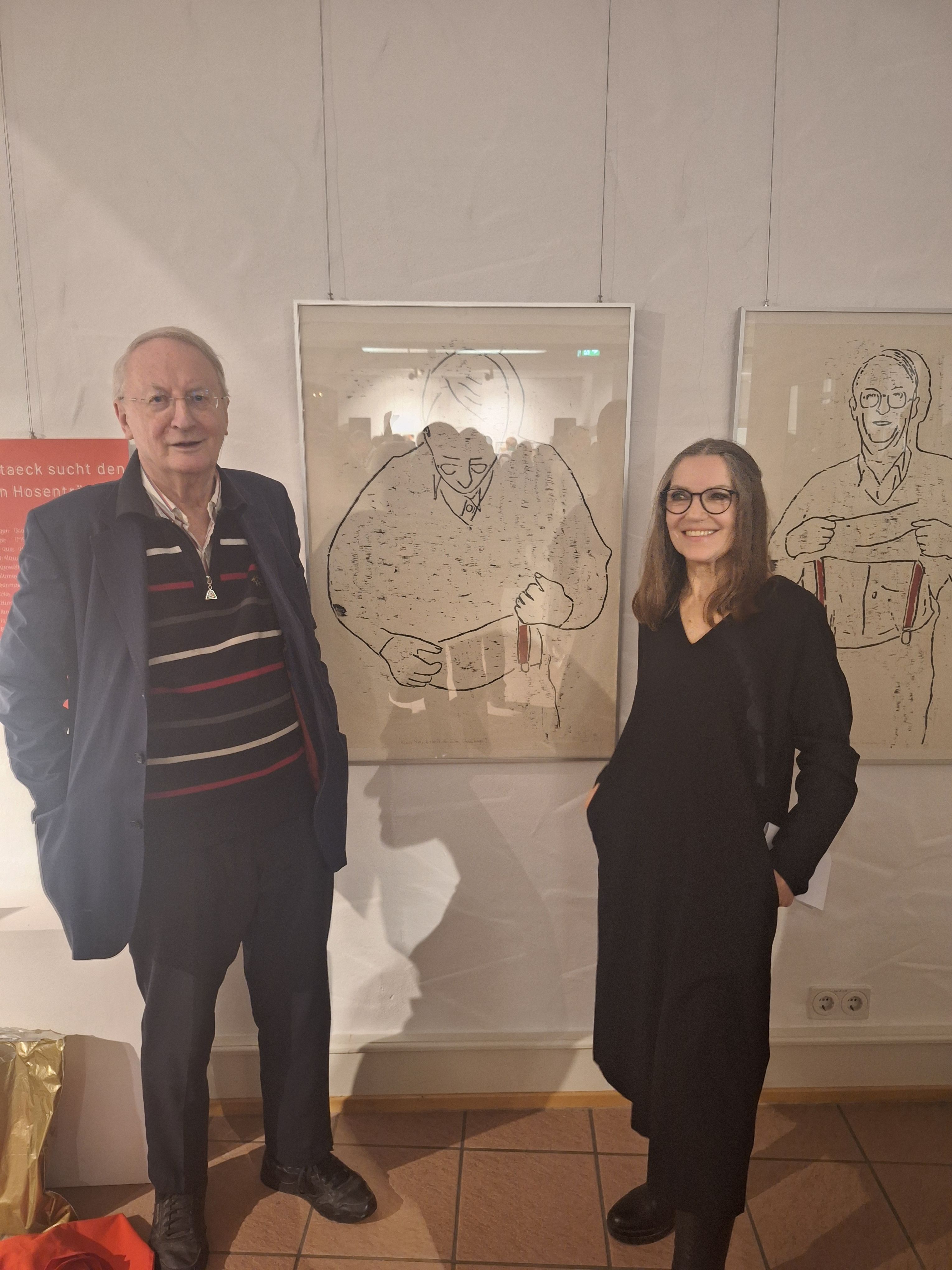 Zwei Personen stehen in einem Ausstellungsraum neben gerahmten Zeichnungen.