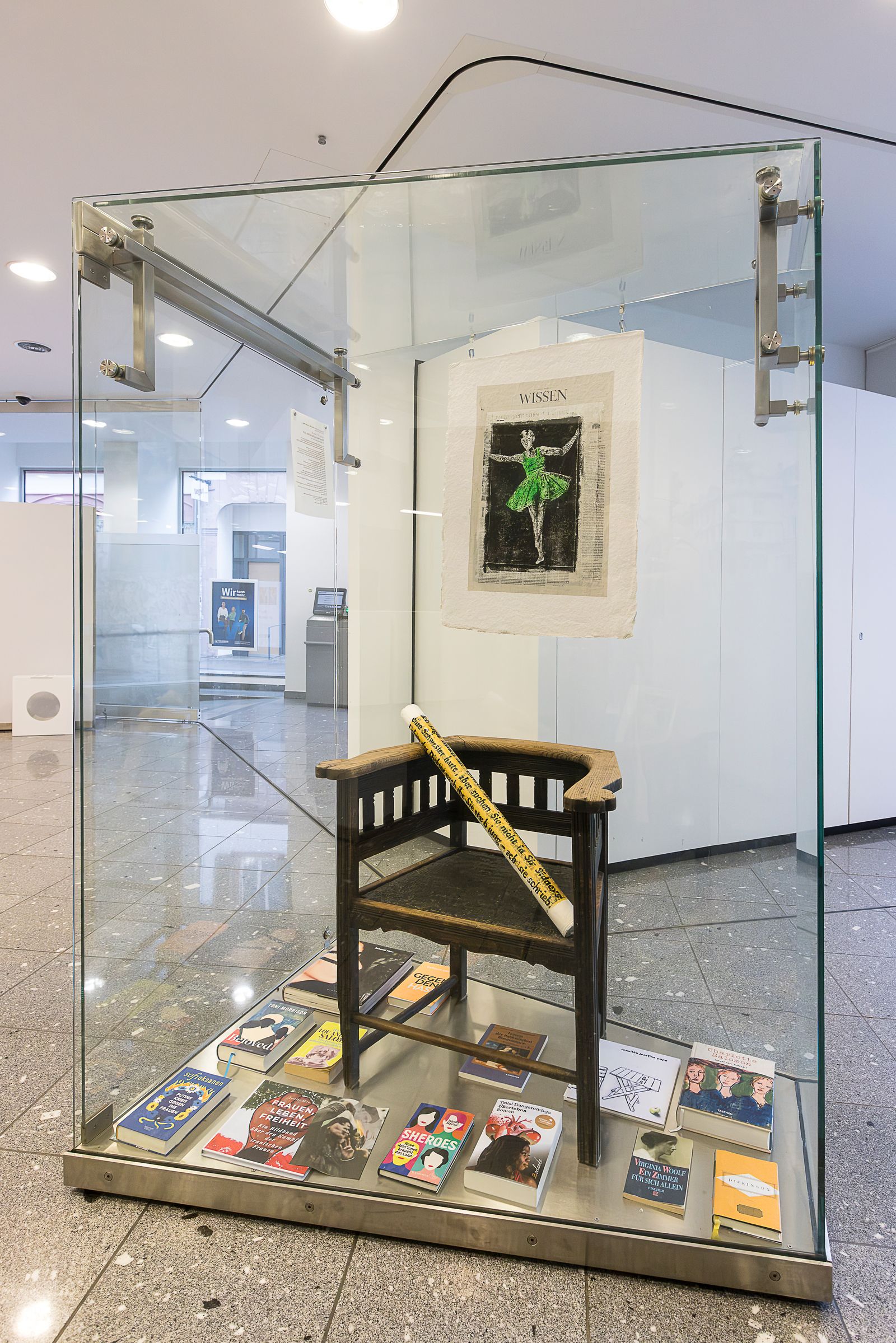 Glasvitrine mit einem Holzstuhl, Büchern und einer hängenden Arbeit mit einer Figur in grünem Kleid.