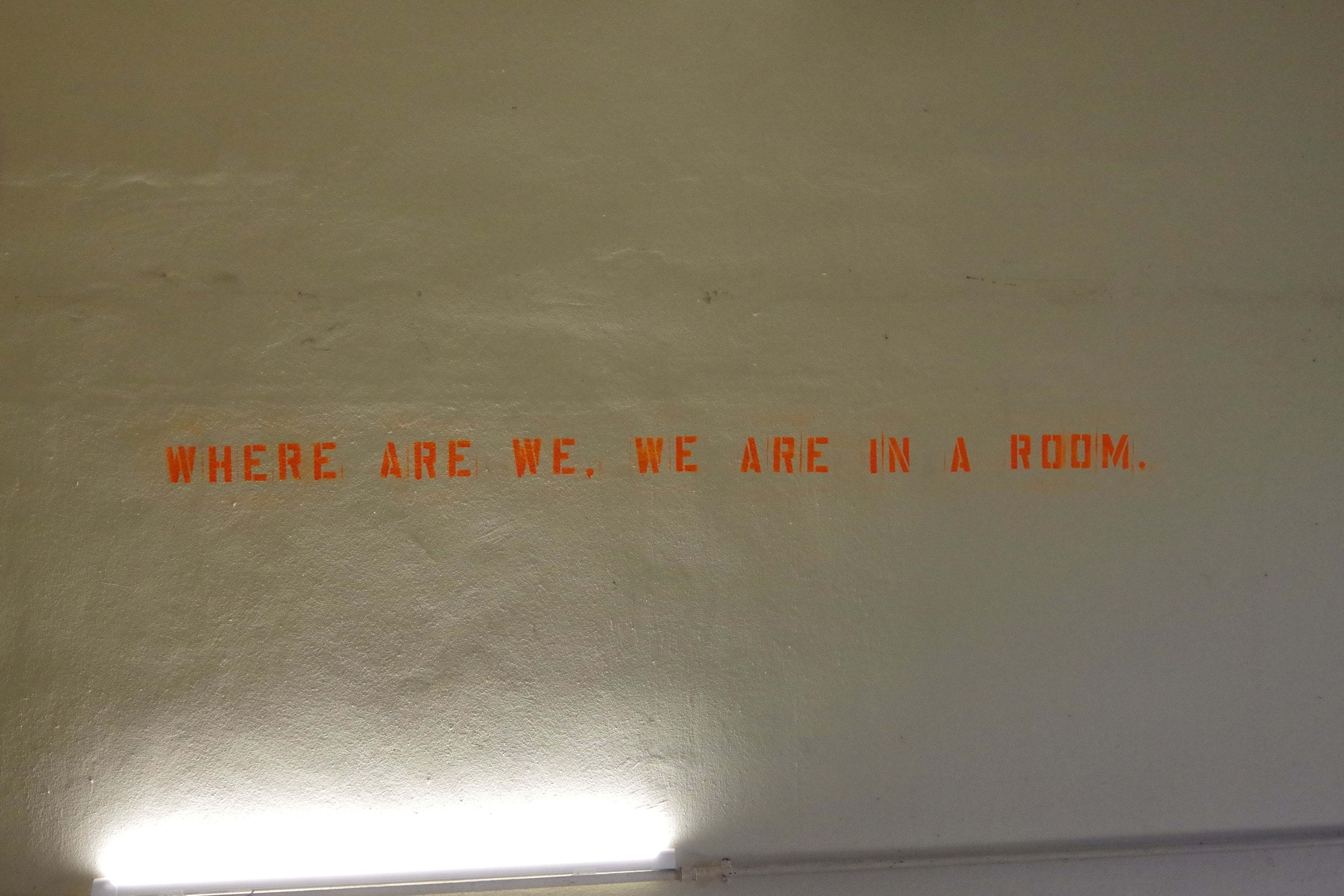 Orangefarbener Schriftzug Where are we, we are in a room an einer grauen Wand.