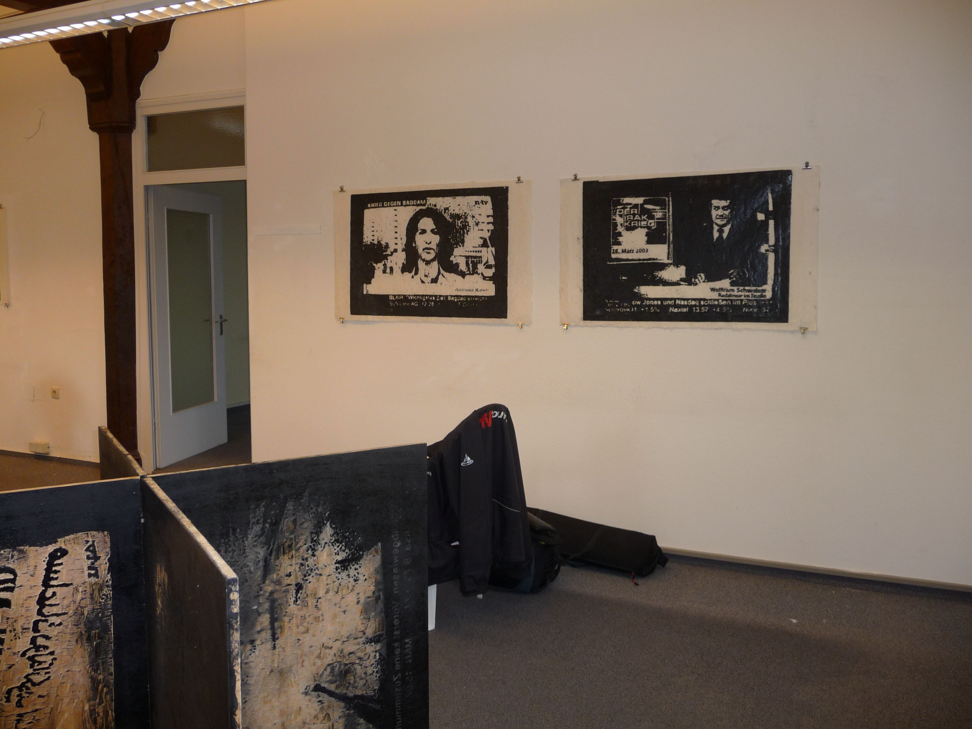 Ausstellungsraum mit zwei gerahmten schwarzweißen Arbeiten und einem dunklen Objekt davor.